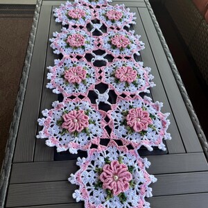 May include: A white and pink crocheted table runner with a floral design. The runner is made with delicate stitches and features pink flowers with white centers.