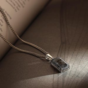 Puede incluir: Un collar de plata con un colgante rectangular. El colgante tiene una piedra oscura y moteada engastada en un marco plateado. La cadena es de diseño delicado. El collar está sobre un libro.