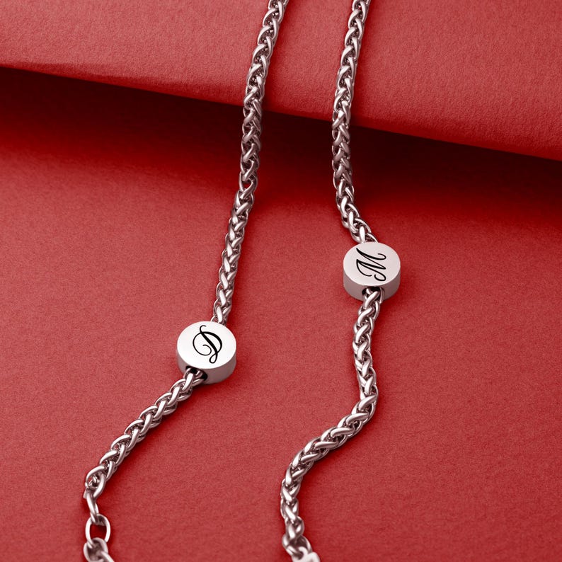May include: A silver-toned necklace with a wheat chain design. The necklace features two circular pendants, each with a black initial. The background is a solid red.