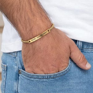 May include: A gold-coloured bracelet featuring a rectangular nameplate engraved with "ADAM - DANIEL - EMMA". The bracelet has a chain-link design. The person is wearing a white shirt and blue jeans.