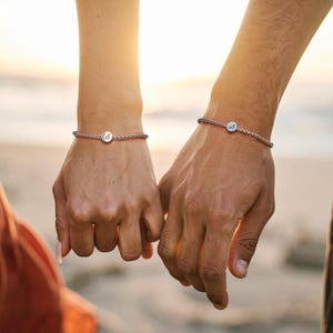 Peut inclure: Deux personnes portant des bracelets en argent assortis avec un petit charme rond sur chacun. Les bracelets sont en cordon tressé avec un accent bleu.