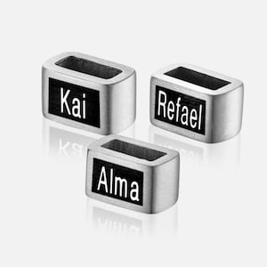May include: Three silver metal nameplate charms with black backgrounds. The charms read "Kai", "Rafael", and "Alma".