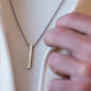 Op de afbeelding: Een zilveren ketting met een zilveren staafhanger met braille-letters.