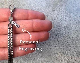 May include: A silver chain necklace with a small silver tag that says "Emma" on it. The tag is attached to the chain with a jump ring. The chain is a herringbone style. The necklace is on a hand, and the text "Personal Engraving" is written below the chain.