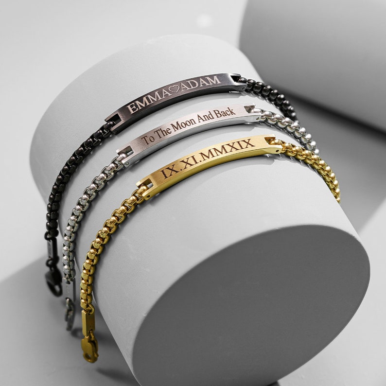 May include: Three bracelets with engraved metal plates. The top bracelet is black with the text "EMMA ADAM" and "To The Moon And Back". The middle bracelet is silver with the text "IX.XI.MMXIX". The bottom bracelet is gold with the text "IX.XI.MMXIX".