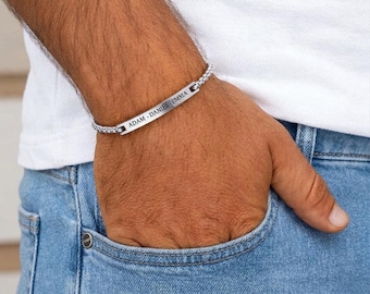 Op de afbeelding: Een zilveren armband met een schakelband en een rechthoekige plaat met de namen "ADAM - DANIEL EMMA" gegraveerd. De armband wordt om een pols gedragen, de persoon draagt een blauwe spijkerbroek en een wit shirt.