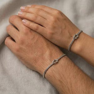 Puede incluir: Dos pulseras plateadas con forma de nudo se muestran en muñecas. Las pulseras tienen un diseño simple y elegante, con un detalle de nudo en el centro. El fondo es una tela neutra de color claro, creando una estética suave y minimalista.