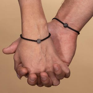 May include: Two people wearing black braided bracelets with small round metal charms. The charms have the letters "R" and "N" engraved on them.