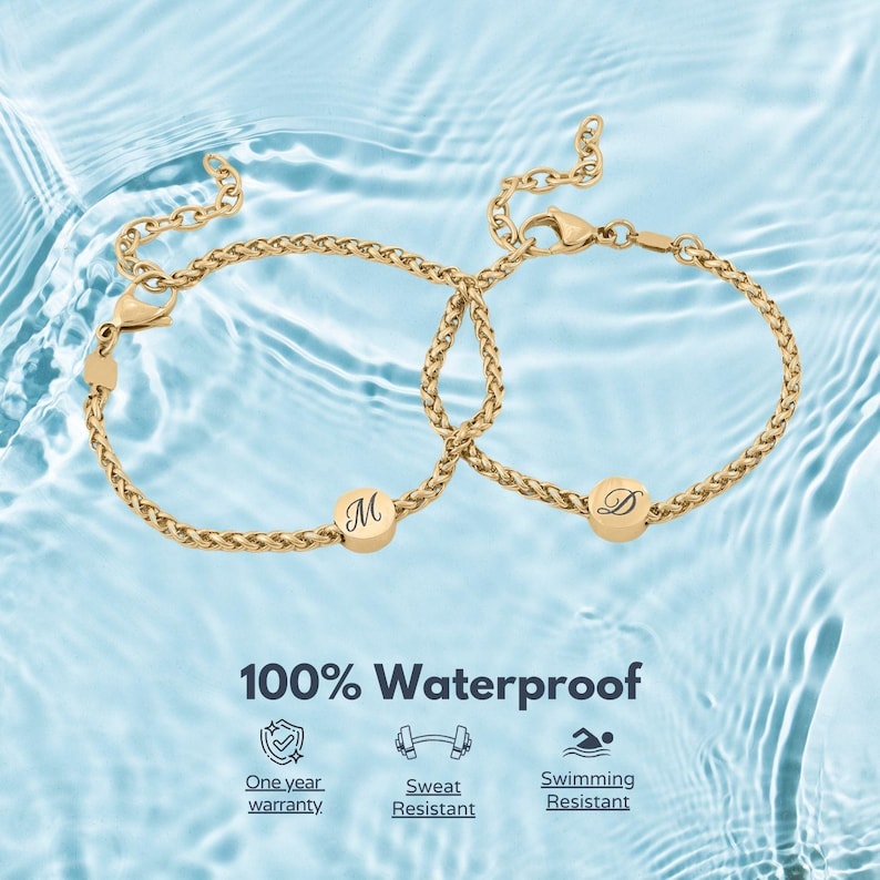 May include: Two gold-colored bracelets with a woven chain design. Each bracelet has a round charm with a letter engraved on it. The bracelets are displayed against a blue water background. The text "100% Waterproof" is visible.