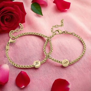 May include: Two gold-colored chain bracelets with initial charms, one with the letter "M" and the other with "D". The bracelets are displayed on a pink fabric with rose petals and a red rose in the background.