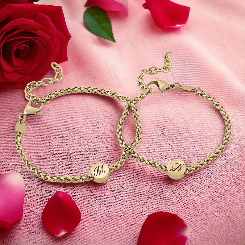 May include: Two gold-colored chain bracelets with initial charms, one with the letter "M" and the other with "D". The bracelets are displayed on a pink fabric with rose petals and a red rose in the background.