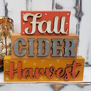 May include: Three wooden blocks stacked on top of each other. The top block is red with white polka dots and the words "Fall" in white. The middle block is brown with white polka dots and the words "Cider" in gray. The bottom block is yellow with white polka dots and the words "Harvest" in orange.