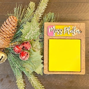 May include: A wooden sign with a yellow sticky note and a pencil-shaped topper that says "Miss Pfeifer". The sign is sitting on a brown wooden surface with a green pine branch and pine cones.