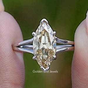 Op de afbeelding: Een verlovingsring met marquise-slijpvorm met een lichtgele edelsteen. De ring heeft een split-schachtontwerp en wordt tussen twee vingers gehouden. De achtergrond is zachtgroen. De tekst "Golden Bird Jewels" is zichtbaar.