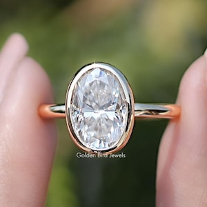 Op de afbeelding: Een gouden ring met een grote, ovaalvormige diamant. De diamant is gezet in een bezel-zetting en de band is van gepolijst goud. De ring wordt tussen twee vingers gehouden tegen een vage groene achtergrond. De tekst "Golden Bird Jewels" is zichtbaar.
