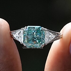 Può includere: Un anello d'argento con una grande pietra preziosa di colore turchese tagliata a forma quadrata, circondata da pietre preziose più piccole e trasparenti. L'anello è su un dito.