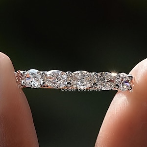 May include: A delicate eternity band featuring a row of oval-shaped diamonds set in a silver band. The band is shown on a finger with pink nail polish.