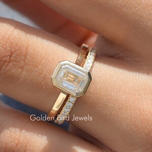 Op de afbeelding: Een gouden ring met een vierkante diamant en een bijpassende band met kleine diamanten. De ring zit aan een vinger.