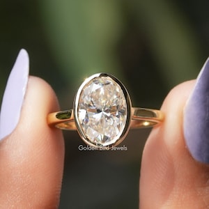 Può includere: Un anello d'oro con un diamante taglio ovale incastonato in una lunetta. La fascia è in oro liscio. L'anello è tenuto tra due dita con smalto viola chiaro. Il testo "Golden Bird Jewels" è visibile.