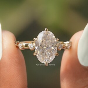 Op de afbeelding: Een gouden verlovingsring met een grote ovale diamant en kleinere diamanten op de band. De ring wordt vastgehouden door een hand met witte nagellak.