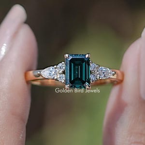 May include: A rose gold engagement ring with a dark green emerald cut gemstone and smaller diamonds on the band.  "Golden Bird Jewels" is printed below the ring.