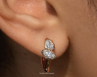 Op de afbeelding: Een close-up van een goudkleurige oorring met twee peervormige, heldere edelstenen. De oorring wordt op een oor gedragen. De merknaam "Golden Bird Jewels" is zichtbaar.