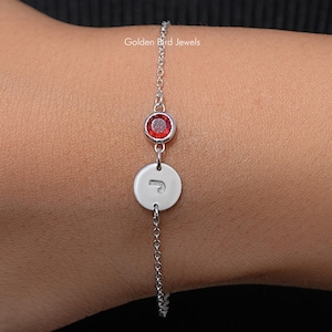 Può includere: Un delicato bracciale a catena color argento caratterizzato da una piccola pietra preziosa rotonda di un rosso vibrante e da un ciondolo circolare in argento inciso con la lettera J. Il gioiello è realizzato con una sottile catena a maglie.