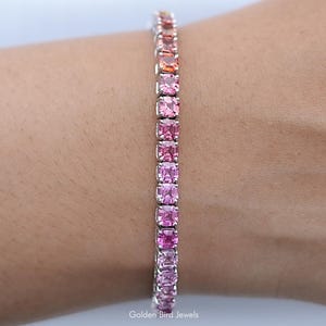 Può includere: Un braccialetto tennis con un gradiente di pietre preziose rosa, che vanno dal rosa chiaro al rosa scuro e all'arancione. Il braccialetto è montato su una fascia in argento o oro bianco. Il braccialetto è al polso di una persona.