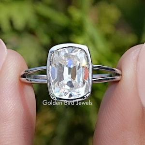 Op de afbeelding: Een zilveren ring met een grote, heldere, kussen geslepen edelsteen. De ring is gezet in een eenvoudige, elegante band. De tekst "Golden Bird Jewels" is zichtbaar onder de ring.