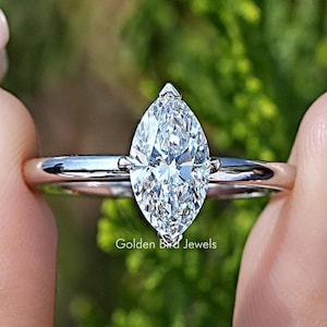 Op de afbeelding: Een verlovingsring met een marquise-geslepen diamant en een dunne, zilveren band. De diamant wordt tussen twee klauwen gehouden en is het middelpunt van de ring. De ring wordt tussen twee vingers gehouden tegen een vage groene achtergrond.