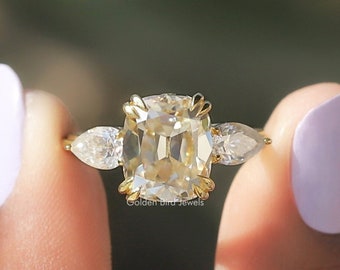 Puede incluir: Un anillo de oro con un gran diamante de talla cojín de color amarillo claro en el centro y dos diamantes más pequeños de talla pera a cada lado.  "Golden Bird Jewels" está impreso debajo del anillo.