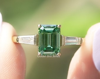 Könnte beinhalten: Ein goldener Ring mit einem grünen Smaragd-Schliff-Edelstein und zwei kleineren Baguette-Schliff-Diamanten auf jeder Seite. "Golden Bird Jewels" ist unter dem Ring gedruckt.