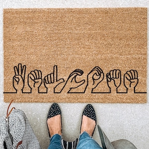 May include: A rectangular doormat made of natural coir fiber. The mat features the word "WELCOME" spelled out in black hand gestures, using American Sign Language. The mat is placed on a concrete surface.