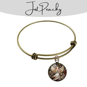 Op de afbeelding: Antiek goudkleurige verstelbare armband met een ronde bedel. De bedel heeft een gedetailleerd engelontwerp met rode, witte en gouden accenten. De armband heeft een eenvoudig, elegant ontwerp.