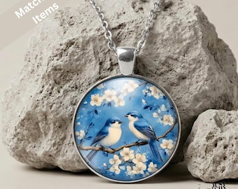 May include: A silver-toned pendant necklace featuring a circular glass cabochon with an illustration of two blue birds perched on a branch surrounded by white cherry blossoms against a blue background, suspended from a delicate silver chain.