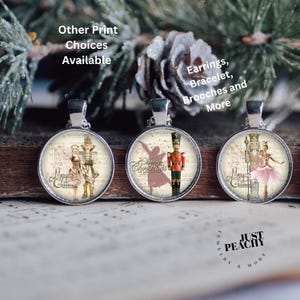 Op de afbeelding: Drie ronde zilveren hangers met kerstthema-ontwerpen. Eén toont een ballerina, een andere een notenkraker en de derde een stel. De hangers staan op een achtergrond van bladmuziek en de tekst "Merry Christmas". De tekst "Other Print Choices Available" is ook zichtbaar.