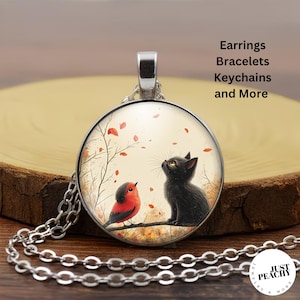 May include: A silver-toned circular pendant necklace featuring an illustration of a black cat and a red bird perched on a branch with falling leaves. The text Earrings Bracelets Keychains and More is visible, along with a Just Peachy logo.