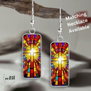 May include: Silver-tone earrings with rectangular pendants featuring a stained-glass cross design. The cross is white, surrounded by a vibrant array of red, yellow, blue, and purple colors. The text "Matching Necklace Available" is visible.