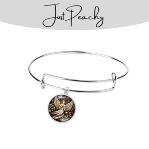 Op de afbeelding: Een verstelbare zilverkleurige armband met een ronde bedel. De bedel toont een gedetailleerde illustratie van een engel met uitgespreide vleugels, tegen een achtergrond van rode, gouden en bruine accenten. De tekst "Just Peachy" staat boven de armband.