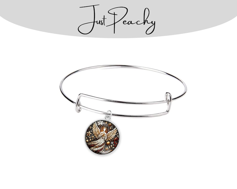 Op de afbeelding: Een verstelbare zilverkleurige armband met een ronde bedel. De bedel toont een gedetailleerde illustratie van een engel met uitgespreide vleugels, tegen een achtergrond van rode, gouden en bruine accenten. De tekst "Just Peachy" staat boven de armband.