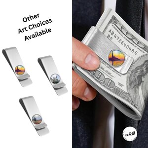 May include: Silver money clips with different art designs. One clip holds a stack of banknotes. The art choices include a sunset scene, the Eiffel Tower, and a windmill scene. The image includes the text "Other Art Choices Available."