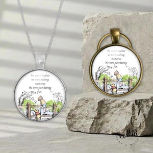 Op de afbeelding: Een zilverkleurige ketting met hanger en een bronskleurige hanger met een ronde illustratie van een kind en een beer bij een houten hek. De tekst luidt: We didn't realize we were making memories. We were just having fun.