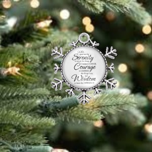 May include: A silver snowflake ornament with a circular center. The center features the words "Serenity," "Courage," and "Wisdom" in elegant script. The ornament is hanging in front of a blurred green Christmas tree.