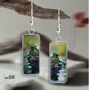 May include: Silver-tone rectangular earrings featuring a green frog in water. The frog has black eyes and is set against a green and brown background. The earrings are suspended from a silver hook. The brand name "JUST PEACHY & MORE" is printed on the left side.