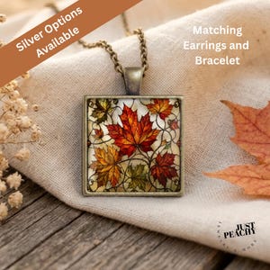 Può includere: Una collana con ciondolo quadrato caratterizzata da un design in stile vetrata con foglie d'acero autunnali nei toni del rosso, arancione e giallo. Il ciondolo ha una cornice in metallo color bronzo e pende da una catena abbinata. Il testo recita Silver Options Available.