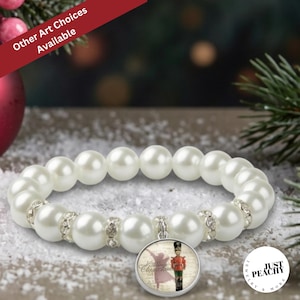Pode incluir: Uma pulseira elástica de pérolas brancas com espaçadores prateados incrustados com cristais transparentes. Um pingente circular pende da pulseira, exibindo um gráfico de um soldado quebra-nozes e o texto "Christmas".