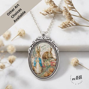 Può includere: Una collana con ciondolo ovale color argento che presenta un'illustrazione dettagliata di due conigli in un giardino, uno dei quali indossa una giacca blu. La cornice ha un bordo decorativo a forma di foglia. Il testo sull'immagine recita Other Art Choices Available e Just Peachy.