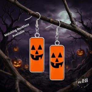 May include: A pair of rectangular drop earrings featuring a bright orange background with a black jack-o'-lantern face design. The earrings hang from silver-toned metal hooks and include the text Matching Necklace Available on the left side.