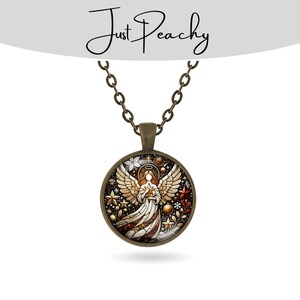 Op de afbeelding: Een ronde hangerketting met een engelontwerp. De hanger heeft een gedetailleerde illustratie van een engel met uitgestrekte vleugels, die een ster vasthoudt. Het ontwerp bevat sterren, bloemen en een rood-wit kleurenschema. De ketting en hanger zijn bronskleurig, met de tekst "Just Peachy" bovenaan.