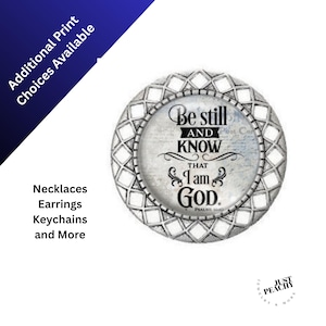 May include: A round, silver-toned pendant with the text "Be still and KNOW that I am GOD" in black script. The pendant has a decorative, geometric border. Additional print choices are available. The image also shows the words "Necklaces, Earrings, Keychains and More."
